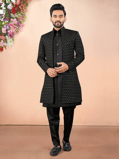 Man wearing a black patterned coat and black pants against a beige background with flowers on the side.