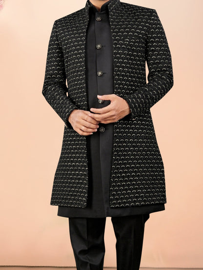 Man wearing a black patterned coat and black pants against a beige background with flowers on the side.