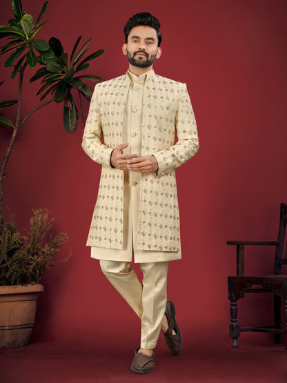 Man wearing a cream sherwani standing against a red background with plants on either side.