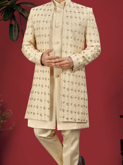 Man wearing a cream sherwani standing against a red background with plants on either side.