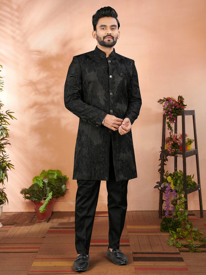 Man wearing a black traditional outfit standing in a decorated room with plants and flowers.