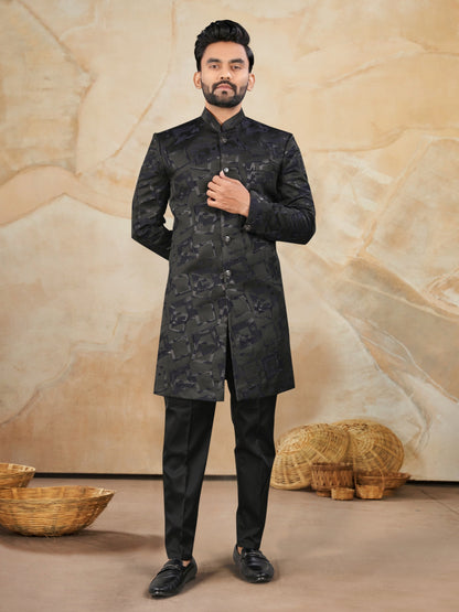 Man wearing a black traditional outfit against a beige textured wall.