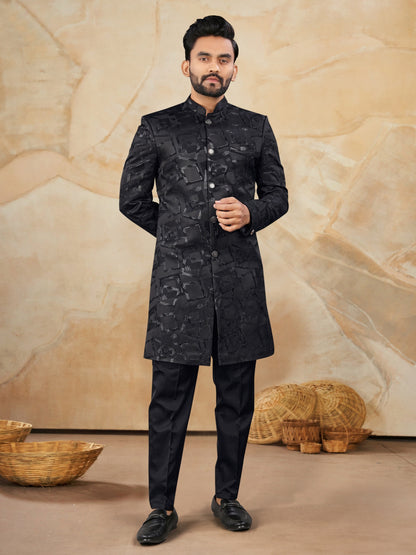 Man wearing a black traditional outfit against a beige textured wall.