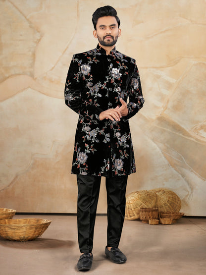 Man wearing a black floral sherwani against a beige textured wall.