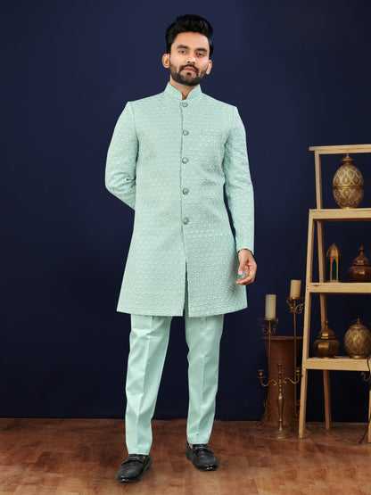 Man wearing a light blue sherwani standing against a dark blue wall with decorative items.