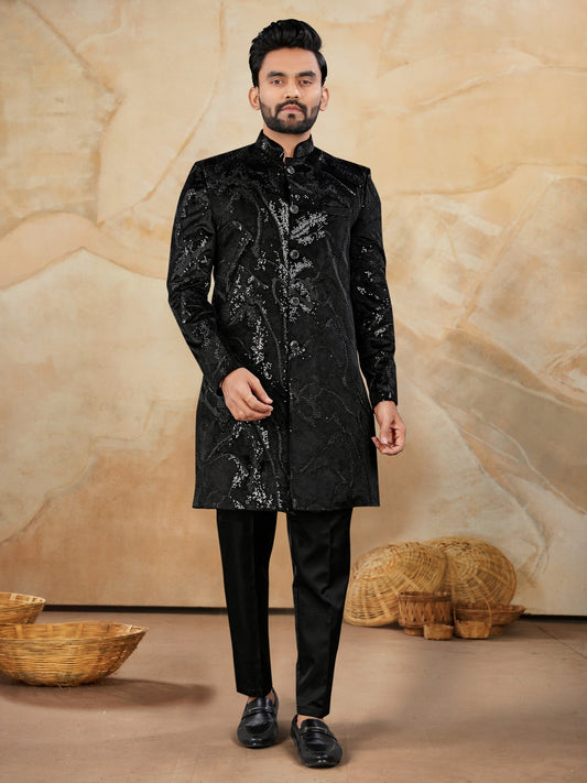 Man wearing a black sherwani with sequin details against a beige textured wall.