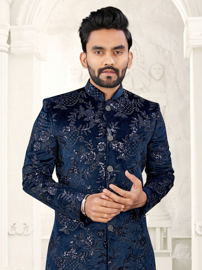 Man wearing a dark blue embroidered sherwani against a white decorative wall.