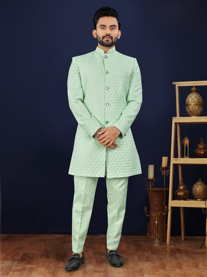 Man wearing a light green traditional suit standing against a dark blue wall with decorative items.