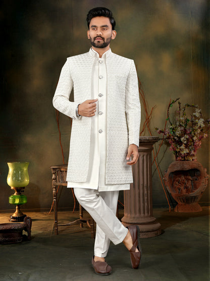 Man wearing a white traditional outfit in a decorative setting with plants and a lamp.