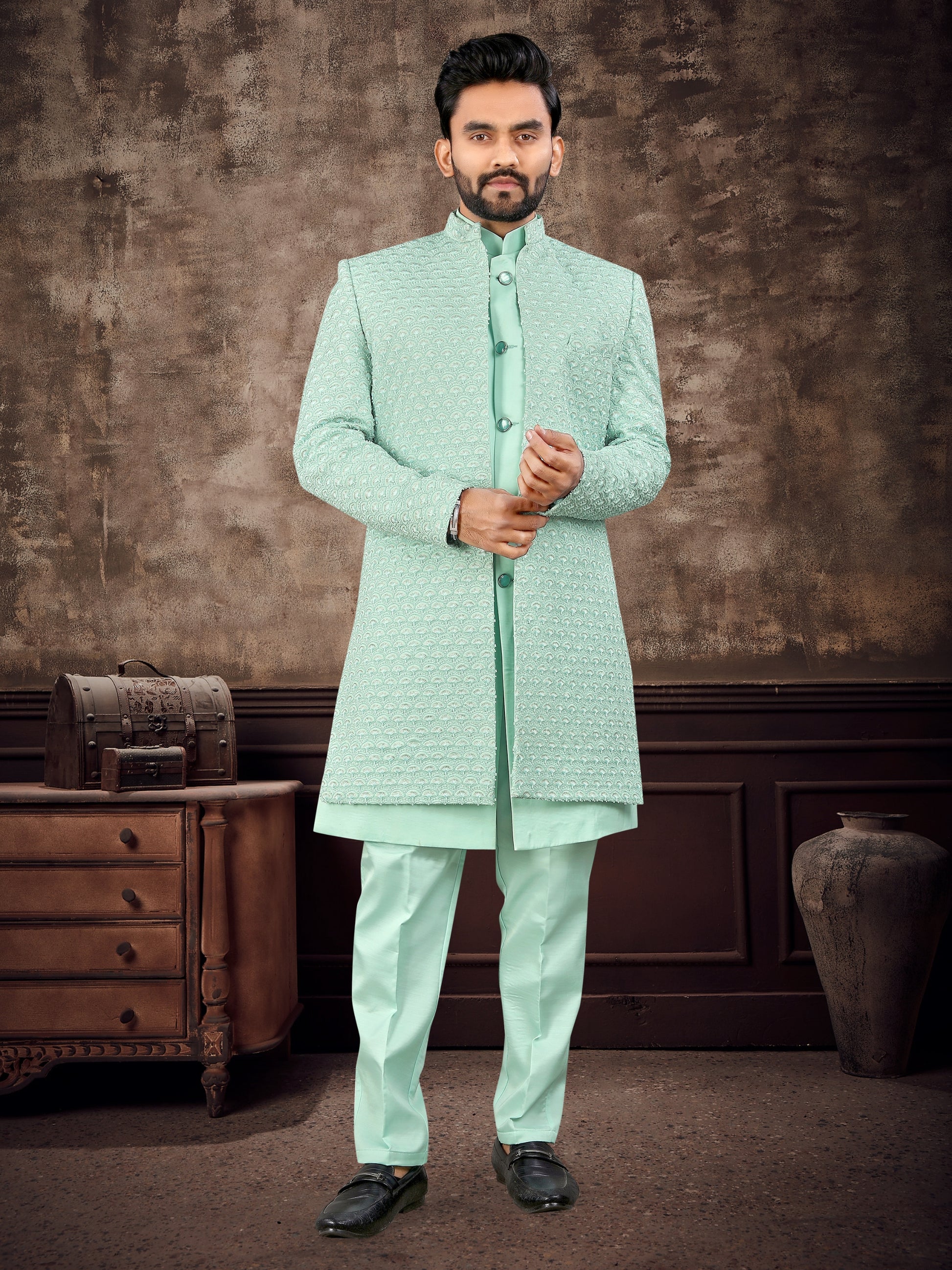 Man wearing a light green traditional outfit against a textured brown wall.