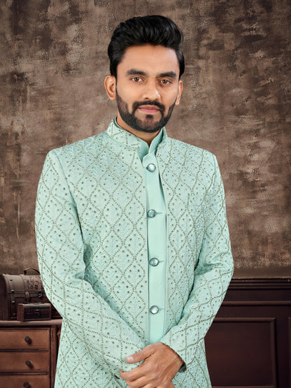 Man wearing a light green traditional outfit against a brown textured wall.