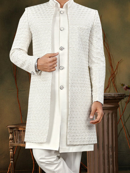 Man wearing a traditional white sherwani in a decorative setting with flowers and a lamp.