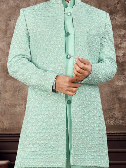 Man wearing a light green traditional outfit against a textured brown wall.