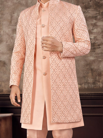 Man wearing a peach sherwani with intricate patterns in a rustic setting.